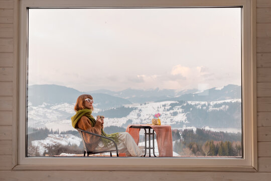 Fototapeta Woman sitting with a drink on terrace of tiny house in the mountains, view through the window from the inside. Concept of small modern cabins for rest and escape to nature
