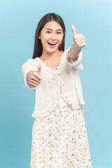 Beautiful young asian woman in white dress with flower pattern making thumbs up sign on both hands isolated on blue background