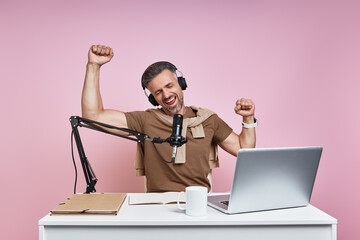 Cheerful man using microphone and gesturing while recording podcast against pink background