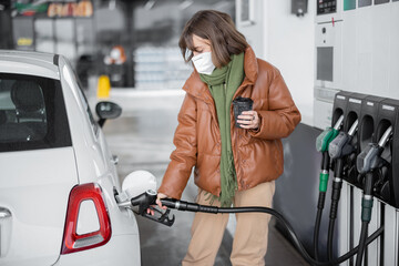 Woman refueling car with gasoline standing with a coffee and holding refueling gun at petrol station. Woman wearing face mask and warm clothes