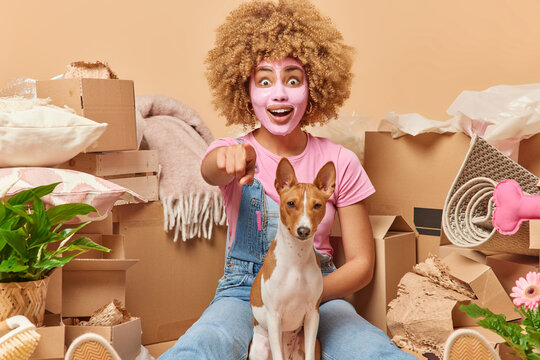 Surprised curly haired woman points directly at camera notices something strange poses with pedigree dog dressed casually appliesbeauty facial mask relocates to new house rents flat for living