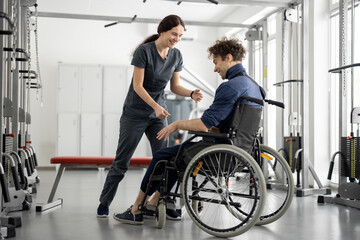 Fototapeta na wymiar Rehabilitation specialist helps a guy stand out of a wheelchair at rehabilitation center. Concept of physical therapy and support for people with disabilities