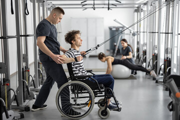Rehabilitation specialist helps a guy in a wheelchair to do exercise on decompression simulator for recovery from injury. Concept of physical therapy for people with disabilities - 526539410