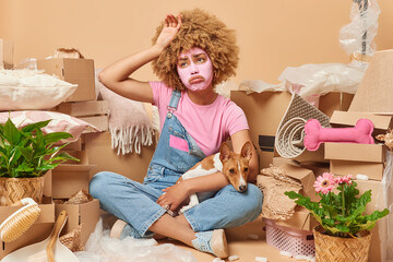 Tired displeased curly haired young woman applies beauty mask on face feels exhausted after relocation and packing personal belongings in cardboard boxes poses with pet on floor in lotus pose