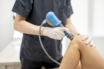 Doctor applies shock wave therapy with special medical equipment on women's knee joints at medical office. Concept of non-invasive technology for treating pain in musculoskeletal system - obrazy, fototapety, plakaty