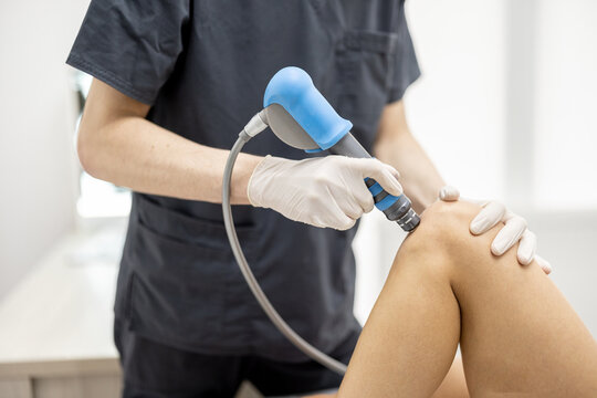 Doctor applies shock wave therapy with special medical equipment on women's knee joints at medical office. Concept of non-invasive technology for treating pain in musculoskeletal system