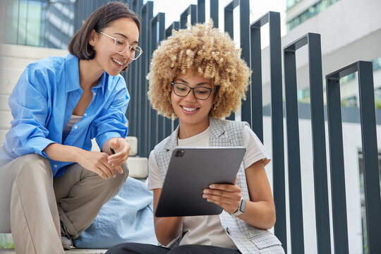Outdoor shot of mixed race millennial girls read some text on digital tablet surf internet search information in internet wear spectacles and stylish clothes have happy expressions. Modern technology