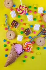 Bright sugar sweets splash from ice cream waffle cone on yellow background. Top view on lollipop, cookies, marshmallow, chocolate, round button