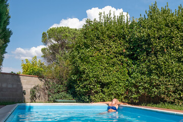 A blonde woman relaxes in the corner of a swimming pool in a garden with many plants