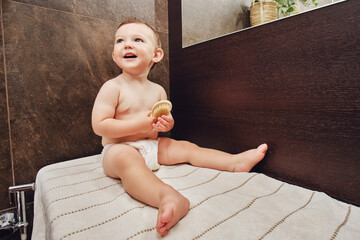 Portrait of a happy toddler baby with a comb in the bathroom by the mirror. Baby boy sitting on a...