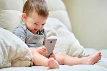 Happy toddler baby boy is sitting with a phone on the sofa in the living room. Child with a...