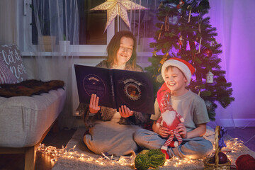 Obraz na płótnie Canvas An adult woman and a boy are reading a book by the Christmas tree in the home living room. Mom and son read fairy tales on New Year Eve