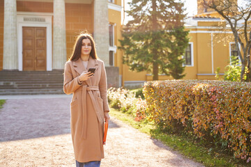Fototapeta premium beautiful woman in a light coat in the sun in the autumn park near a beautiful building and low yellow bushes