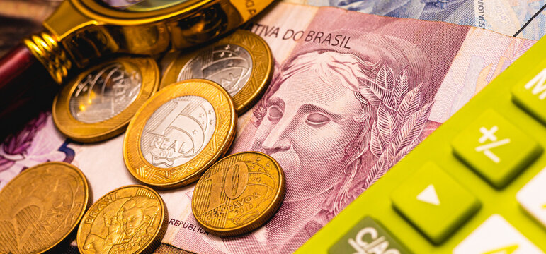 Notas do Real Brasileiro e moedas em tempo de crise financeira no Brasil. Uma lupa e uma calculadora na composição da imagem. Economia brasileira, finanças e a inflação.