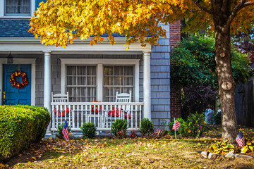 Blue shinngled two stor house entrance with porch and rocking chairs and American flags and a fall...