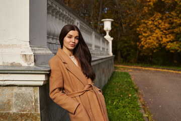 Naklejka premium a beautiful woman in a brown coat smiles and stands leaning against white columns in an autumn park