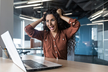 Critical situation at work business woman in shock screams and tears her hair on her head, disappointed Latin American woman looks at the laptop screen, female employee works inside the office