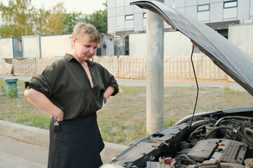 Confident blonde woman stands in front of a car with the hood up. Woman waiting for help on the road. Woman in a black blouse stands next to a broken down car.