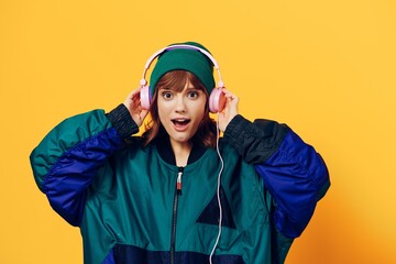 a happy, joyful woman in a green jacket and hat enjoys music standing in pink headphones on a yellow background