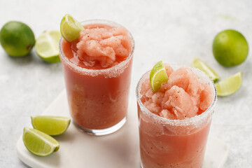 Two glasses with tall long watermelon drink with crushed ice, sugar rims, lime slices on marble board on grey background