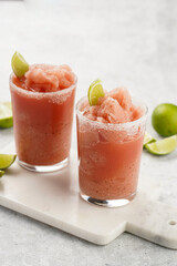 Two glasses with tall long watermelon drink with crushed ice, sugar rims, lime slices on marble board on grey background