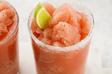 Two glasses with tall long watermelon drink with crushed ice, sugar rims, lime slices on marble...