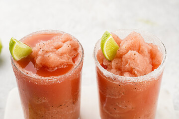 Two glasses with tall long watermelon drink with crushed ice, sugar rims, lime slices on marble board on grey background, close-up
