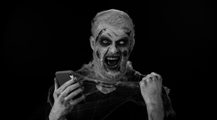 Happy excited sinister man in carnival costume of Halloween crazy zombie looking smartphone display sincerely rejoicing win, receiving message with good news, success luck. Cosplay wounded undead