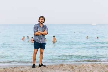 A handsome, young hippie man in a shirt and shorts stands on the beach on the sand against the backdrop of the sea, ocean and people swimming in the summer season.