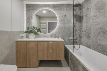 Modern minimalist bathroom interior design with wooden furniture, grey stone tiles, round mirror.  Aesthetic simple interior design concept.