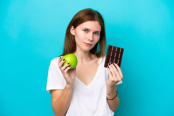 Young English woman isolated on blue background taking a chocolate tablet in one hand and an apple...