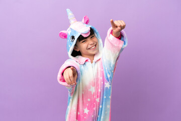 Little kid wearing a unicorn pajama isolated on purple background points finger at you while smiling