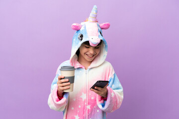 Little kid wearing a unicorn pajama isolated on purple background holding coffee to take away and a mobile