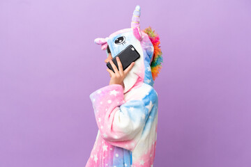 Little kid wearing a unicorn pajama isolated on purple background keeping a conversation with the mobile phone with someone