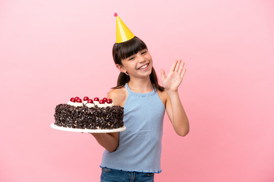 Little caucasian kid holding birthday cake isolated in pink background saluting with hand with happy expression