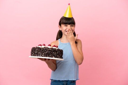 Little caucasian kid holding birthday cake isolated in pink background happy and smiling covering mouth with hand