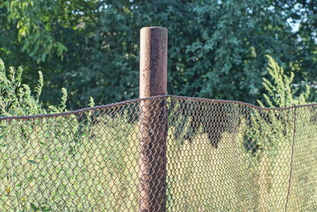 Fototapeta premium part of a fence wall made of brown rusty metal mesh on an iron post on a rural street against the backdrop of green vegetation