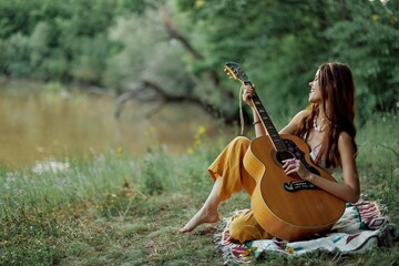 A hippie woman playing her guitar smiles and sings songs in nature sitting on a plaid in the...