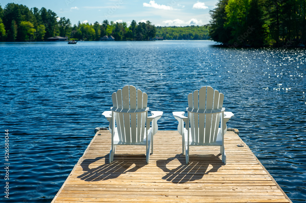 Wall mural beautiful sunny morning, two white adirondack chairs on a cottage wooden dock facing the blue water 