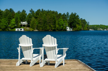Two white Adirondack chairs on a wooden dock facing the blue water of a lake in Muskoka, Ontario Canada. Cottages are visible across the water nestled between green trees.