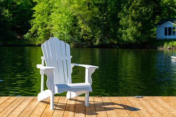 Single white Adirondack chair sitting on a wooden dock in Muskoka Ontario with a calm lake in...