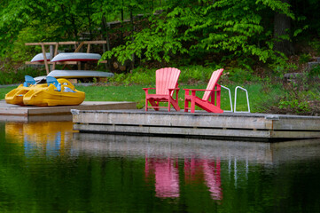 Two red Adirondack chairs on a cottage wookend dock reflecting on the waters of a calm lake in Muskoka. In background a pedal boat and some canoes are visible.