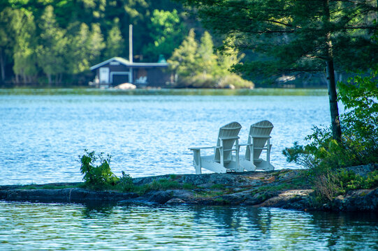 Two white Adirondack chairs on a rock formation facing the blue water of a lake in Muskoka, Ontario Canada. A cottage is visible in background.