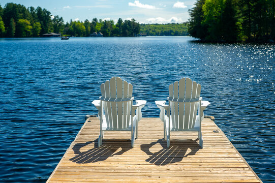 Beautiful sunny morning, two white Adirondack chairs on a cottage wooden dock facing the blue water of a lake in Muskoka, Ontario Canada.