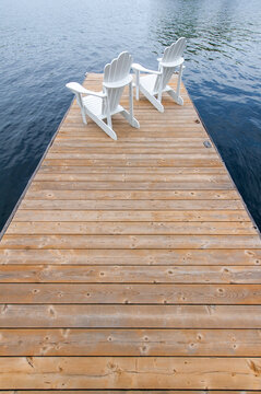 Two white Adirondack chairs on a cottage wooden dock facing the blue water of a lake in Muskoka, Ontario Canada.