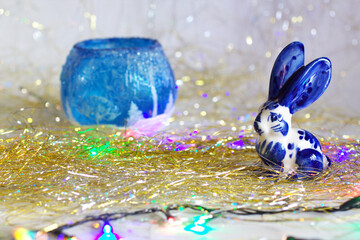 Selective focus of a cute rabbit on a golden tinsel. A symbol of new year 2023 with a candle holder and colorful garland. Christmas, celebration, decorations, winter holidays concept background