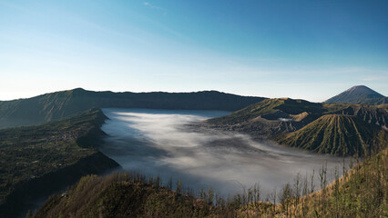 mystic and surreal panoramic mountain view of Bromo volcano craters with foggy surface and green vegetation including Cemoro Lawang village during morning sunshine