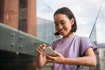 Smiling asian woman using mobile phone listening music standing on the street. Happy female wearing casual clothing holding smartphone shopping online, playing mobile game 
