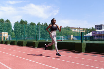 Obraz premium Young African American athlete with long braids runs on red track of city stadium preparing for competitions. Fit black woman in sportswear trains hard to win upcoming marathon on hot summer day