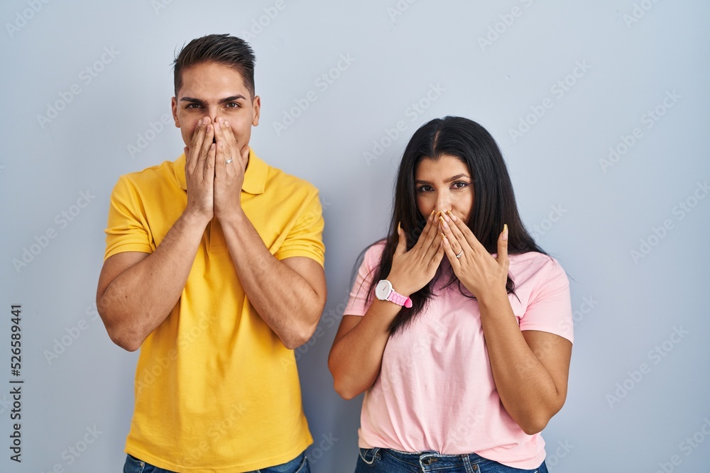 Poster young couple standing over isolated background laughing and embarrassed giggle covering mouth with h